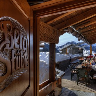 © Bergstüble | Michaela Beck Dornach A cozy inn with wooden decorations and a view of snow-covered mountains. Outside, guests are sitting at a table and enjoying the alpine atmosphere. | © Bergstüble | Michaela Beck Dornach