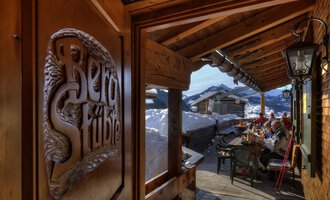 © Bergstüble | Michaela Beck Dornach A cozy inn with wooden decorations and a view of snow-covered mountains. Outside, guests are sitting at a table and enjoying the alpine atmosphere. | © Bergstüble | Michaela Beck Dornach