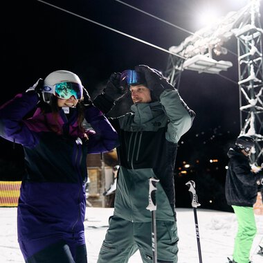 A pair of skiers is preparing for night skiing. In the background, other skiers and a lit ski lift can be seen. | © Kleinwalsertal Tourismus | Oliver Farys