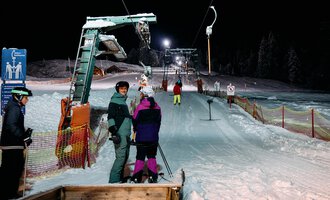 Skiers are waiting at an illuminated ski lift at night. The slopes are covered in snow and surrounded by trees. | © Kleinwalsertal Tourismus | Oliver Farys
