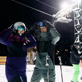 © Kleinwalsertal Tourismus | Oliver Farys Ein Paar Skifahrer bereitet sich auf das Skifahren bei Nacht vor. Im Hintergrund sind weitere Skifahrer und eine beleuchtete Skiliftanlage zu sehen. | © Kleinwalsertal Tourismus | Oliver Farys
