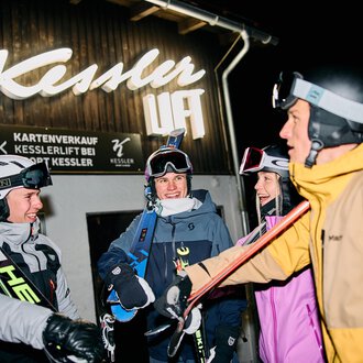 © Kleinwalsertal Tourismus | Oliver Farys A pair of skiers is preparing for night skiing. In the background, other skiers and a lit ski lift can be seen. | © Kleinwalsertal Tourismus | Oliver Farys