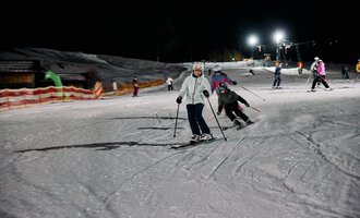 © Kleinwalsertal Tourismus | Oliver Farys Skiers are waiting at an illuminated ski lift at night. The slopes are covered in snow and surrounded by trees. | © Kleinwalsertal Tourismus | Oliver Farys