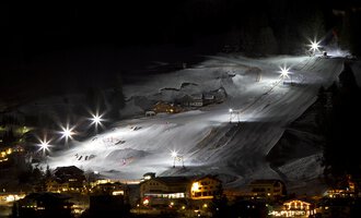 © Sport Kessler GmbH A lit ski slope at night with several skiers. In the foreground, huts and lights are visible. | © Sport Kessler GmbH