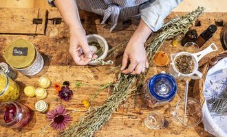 A person is preparing herbs and flowering plants on a wooden table. Next to them are various glass containers with ingredients and utensils for processing. | © Kleinwalsertal Tourismus - Bettina Kessler