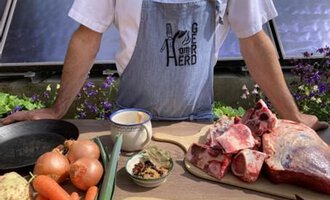 A cook stands at a table with fresh ingredients, including vegetables and meat. In the background, flowers and a bright environment are visible. | © Gerd am Herd | Gerd Hammerer