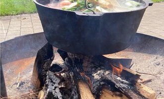 A cauldron with fresh ingredients over an open fire. In the background, green hills and trees can be seen. | © Gerd am Herd | Gerd Hammerer