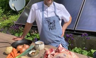 A chef stands at a wooden table outdoors and smiles. In front of him are various fresh ingredients and a large piece of meat. | © Gerd am Herd | Gerd Hammerer