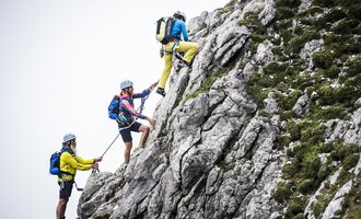 Three climbers are ascending a steep rock. They are wearing helmets and climbing gear as they securely hold on to ropes. | © Bergschule Kleinwalsertal | Dominik Berchtold