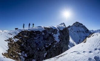 © Kleinwalsertal Tourismus eGen | Dominik Berchtold A group of hikers stands on a snowy summit. The sun shines brightly over the mountains. | © Kleinwalsertal Tourismus eGen | Dominik Berchtold