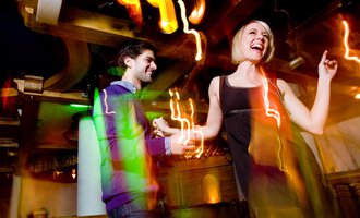 © Aparthotel Kleinwalsertal Haller GmbH & Co KG A couple dances happily in a lively club. Colorful lights and a festive atmosphere surround them. | © Aparthotel Kleinwalsertal Haller GmbH & Co KG