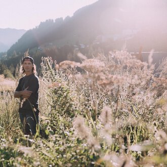 © Das Naturhotel Chesa Valisa****s A young man stands in a blooming field with tall grasses. The sun is shining and the mountains are visible in the background. | © Das Naturhotel Chesa Valisa****s