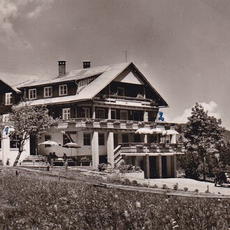 Ein historisches Hotel in den Bergen, umgeben von Bäumen und Wiesen. Im Vordergrund sind alte Autos und eine Terrasse mit Sonnenschirmen zu sehen. | © Genuss- & Aktivhotel Sonnenburg
