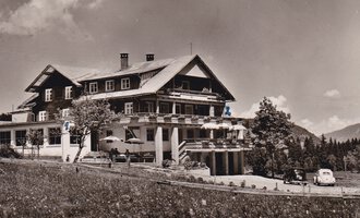Ein historisches Hotel in den Bergen, umgeben von Bäumen und Wiesen. Im Vordergrund sind alte Autos und eine Terrasse mit Sonnenschirmen zu sehen. | © Genuss- & Aktivhotel Sonnenburg