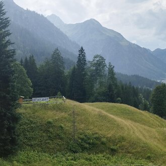 © Nicole Lughammer A picturesque landscape with green hills and tall mountains. In the foreground, there is a small cabin surrounded by trees. | © Nicole Lughammer
