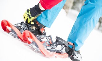 © Kleinwalsertal Tourismus eGen | Oliver Farys A person straps on snowshoes while standing in the snow. The ground is white and the clothing is colorful. | © Kleinwalsertal Tourismus eGen | Oliver Farys