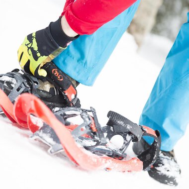 © Kleinwalsertal Tourismus eGen | Oliver Farys A person straps on snowshoes while standing in the snow. The ground is white and the clothing is colorful. | © Kleinwalsertal Tourismus eGen | Oliver Farys
