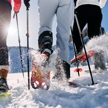© Kleinwalsertal Tourismus | Oliver Farys Three people are hiking in the snow with snowshoes. The sun is shining and snow is spraying from their movements. | © Kleinwalsertal Tourismus | Oliver Farys