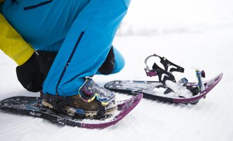 Eine Person zieht Schneeschuhe an, während sie auf einem verschneiten Boden sitzt. Die Ausrüstung und Kleidung sind für winterliche Outdoor-Aktivitäten geeignet. | © Kleinwalsertal Tourismus | Oliver Farys
