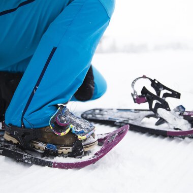 Eine Person zieht Schneeschuhe an, während sie auf einem verschneiten Boden sitzt. Die Ausrüstung und Kleidung sind für winterliche Outdoor-Aktivitäten geeignet. | © Kleinwalsertal Tourismus | Oliver Farys