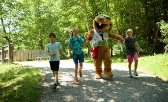 A cheerful group of children is running along a path in the forest with a mascot. The sun is shining through the trees, creating a relaxed atmosphere. | © Kleinwalsertal Tourismus | Oliver Farys