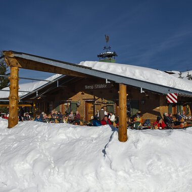 © Bergstüble | Michaela Beck Dornach A cozy mountain cabin in the snow with guests on the terrace. The sky is clear and blue, and the surroundings are wintry. | © Bergstüble | Michaela Beck Dornach