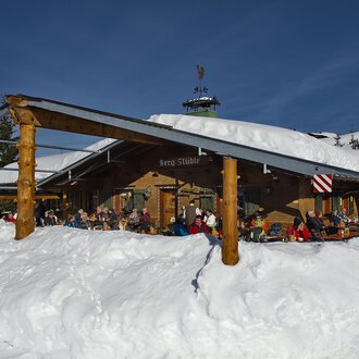 © Bergstüble | Michaela Beck Dornach Eine gemütliche Berghütte im Schnee mit Gästen auf der Terrasse. Der Himmel ist klar und blau, und die Umgebung ist winterlich. | © Bergstüble | Michaela Beck Dornach