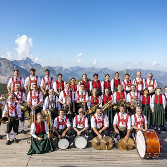 © Trachtenkapelle Riezlern A brass band in traditional attire stands on a platform in the mountains. Impressive mountains and a blue sky are visible in the background. | © Trachtenkapelle Riezlern