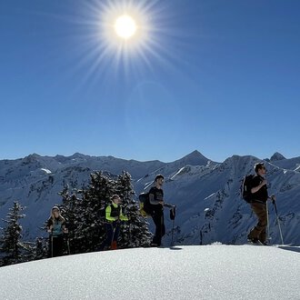A group of hikers stands on a snow-covered summit under bright sunlight. In the background, snow-covered mountains can be seen. | © Abenteuer Vertical | Christian Kohler