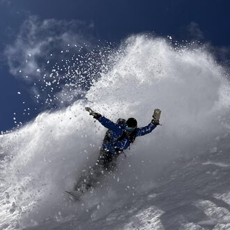 © Abenteuer Vertical | Christian Kohler A snowboarder rides through fresh snow, creating a spray cloud. The sky is clear and blue, ideal for winter sports. | © Abenteuer Vertical | Christian Kohler
