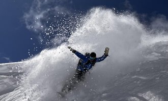 © Abenteuer Vertical | Christian Kohler A snowboarder rides through fresh snow, creating a spray cloud. The sky is clear and blue, ideal for winter sports. | © Abenteuer Vertical | Christian Kohler