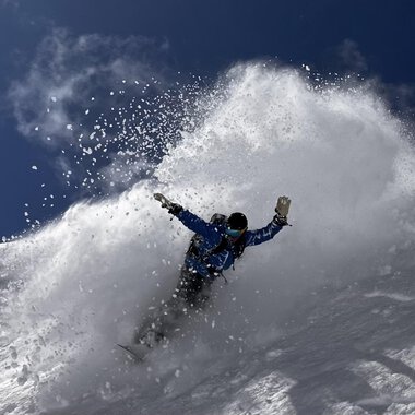© Abenteuer Vertical | Christian Kohler A snowboarder rides through fresh snow, creating a spray cloud. The sky is clear and blue, ideal for winter sports. | © Abenteuer Vertical | Christian Kohler