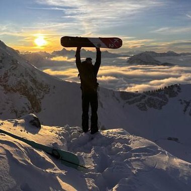 © Abenteuer Vertical | Christian Kohler A snow sports enthusiast stands on a snowy peak, holding a snowboard up high. In the background, the sun shines between the mountains and a blanket of clouds glows in golden colors. | © Abenteuer Vertical | Christian Kohler