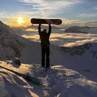 © Abenteuer Vertical | Christian Kohler A snow sports enthusiast stands on a snowy peak, holding a snowboard up high. In the background, the sun shines between the mountains and a blanket of clouds glows in golden colors. | © Abenteuer Vertical | Christian Kohler