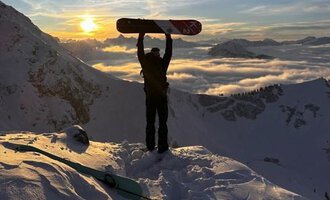 © Abenteuer Vertical | Christian Kohler A snow sports enthusiast stands on a snowy peak, holding a snowboard up high. In the background, the sun shines between the mountains and a blanket of clouds glows in golden colors. | © Abenteuer Vertical | Christian Kohler