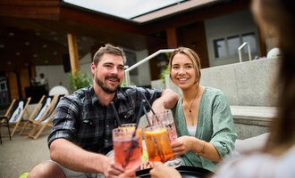 A cheerful couple enjoying cocktails together. Cozy seating is visible in the background. | © Kleinwalsertal Tourismus | Oliver Farys