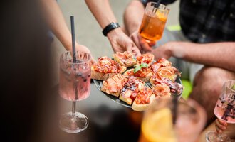 A tray with antipasti, garnished with ham and tomatoes. Surrounded by drinks and people enjoying a sociable time. | © Kleinwalsertal Tourismus | Oliver Farys