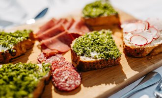A pretty wooden board with various open-faced sandwiches. The snacks are decorated with green herbs, ham, and radishes. | © Kleinwalsertal Tourismus eGen | Oliver Farys