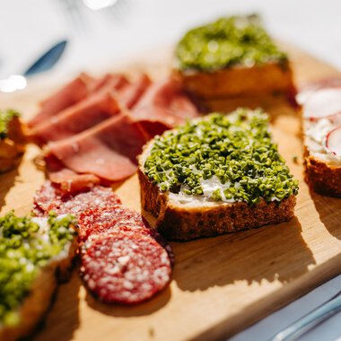 A pretty wooden board with various open-faced sandwiches. The snacks are decorated with green herbs, ham, and radishes. | © Kleinwalsertal Tourismus eGen | Oliver Farys