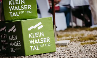Green dice with the inscription "Klein Walsertal since 1302". In the background, chairs and tables in an outdoor area are visible. | © Kleinwalsertal Tourismus eGen | Oliver Farys