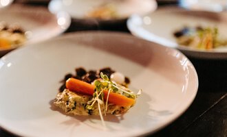 A beautifully arranged plate with a small portion of vegetables and a carrot. In the background, more plates can be seen. | © Kleinwalsertal Tourismus eGen | Oliver Farys