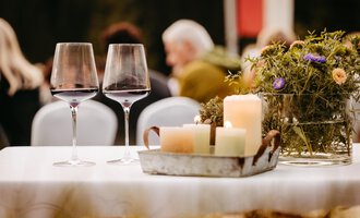 A beautifully set table with two wine glasses and rustic decor. In the background, guests can be seen at an event. | © Kleinwalsertal Tourismus eGen | Oliver Farys