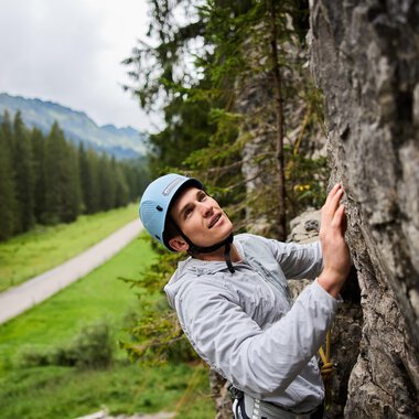 A climber working on a rock face is wearing a helmet and concentrating on his ascent. In the background, trees and a road can be seen in a green landscape. | © Bergschule Kleinwalsertal | Oliver Farys