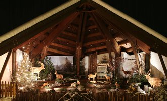 A festive setting with a wooden structure and illuminated animals. In the background, Christmas trees and a winter landscape can be seen. | © Kleinwalsertal Tourismus | Frank Drechsel