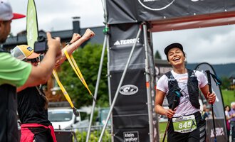 A triathlete runs happily over the finish line and is celebrated by her supporters. In the background, the event banners and a green landscape can be seen. | © Kleinwalsertal Tourismus - Dominik Berchtold