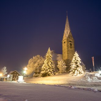 © Kleinwalsertal Tourismus | Dominik Berchtold A snowy landscape at night with a church and illuminated trees. The sky is dark blue and the road is visible. | © Kleinwalsertal Tourismus | Dominik Berchtold