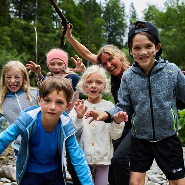 A group of children and an adult standing at the riverbank. They are having fun and posing with happy facial expressions. | © Kleinwalsertal Tourismus | Oliver Farys