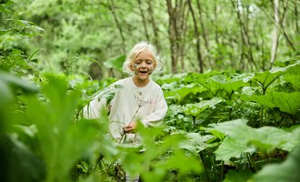 A girl with blonde hair plays happily in a green forest. Surrounded by large plants, she radiates joy and carefreeness. | © Kleinwalsertal Tourismus | Oliver Farys