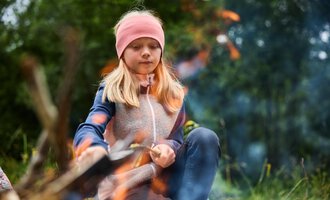 A girl sits by the campfire, focusing intently on the flames. The background shows a green meadow landscape. | © Kleinwalsertal Tourismus | Oliver Farys