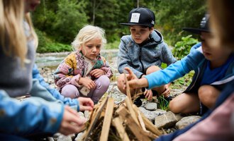 A group of children gathers around a small campfire. They are surrounded by nature and focused on making the fire. | © Kleinwalsertal Tourismus | Oliver Farys
